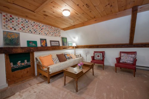 Cozy attic living space with wooden beams, a sofa, two red chairs, and colorful artwork on the walls.