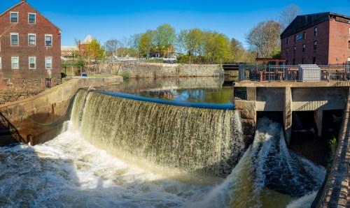 A scenic view of a waterfall cascading over a dam, surrounded by greenery and historic brick buildings.