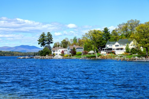 Scenic lakeside view with charming houses, trees, and mountains under a clear blue sky.
