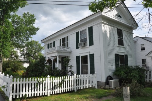 A white two-story house with green shutters, surrounded by a white picket fence and greenery, under a cloudy sky.