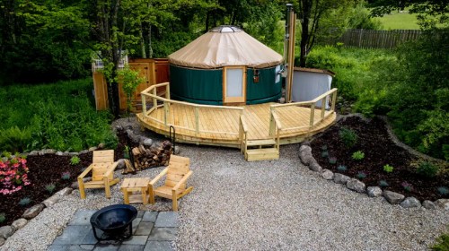 A cozy yurt surrounded by greenery, with a wooden deck, fire pit, and seating area in a landscaped garden.