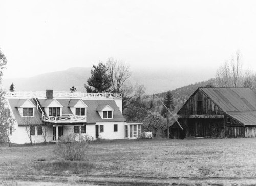 A black and white photo of a farmhouse with a barn, set against a mountainous landscape and cloudy sky.