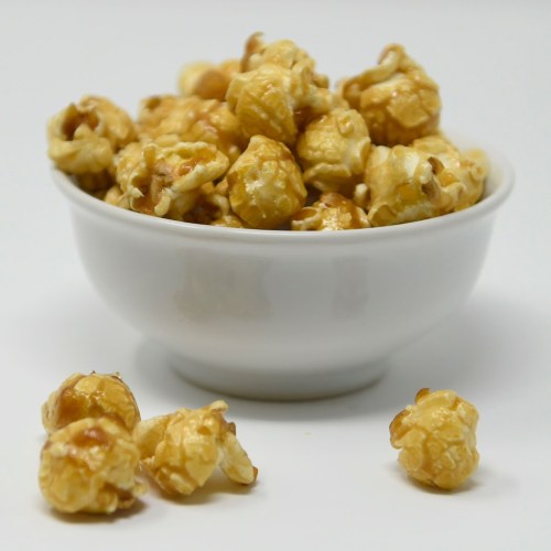 A white bowl filled with caramel popcorn, with a few pieces scattered around it on a plain background.
