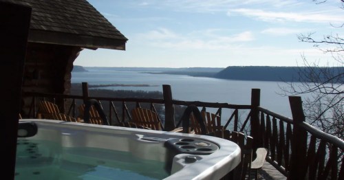 A hot tub on a deck overlooking a serene lake and distant hills under a clear blue sky.