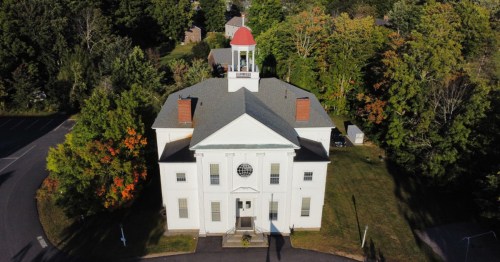 Aerial view of a white building with a red roof and cupola, surrounded by trees in autumn colors.
