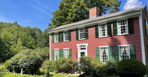 A charming red house with green shutters surrounded by lush greenery and trees under a clear blue sky.
