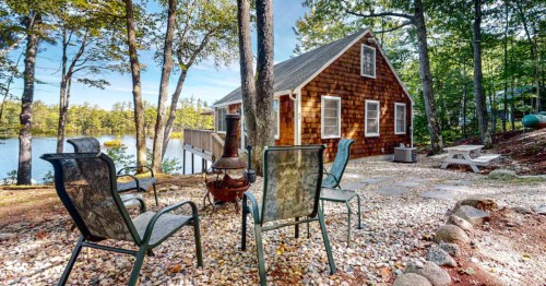 A cozy cabin by a lake, surrounded by trees, with outdoor seating and a fire pit on a pebbled patio.