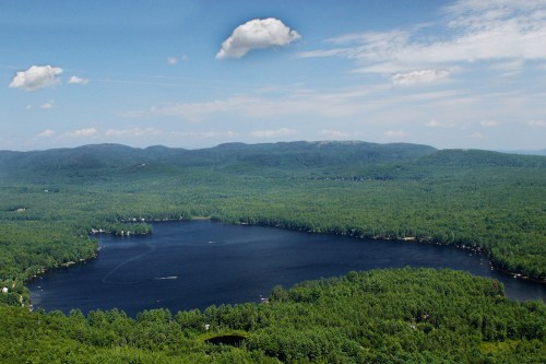 Aerial view of a serene lake surrounded by lush green forests and distant mountains under a blue sky with clouds.