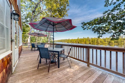 A wooden deck with tables and umbrellas overlooking a calm lake surrounded by trees under a clear blue sky.