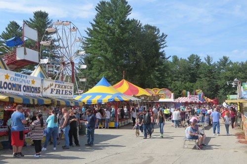 A bustling fairground with colorful tents, food stalls, and a Ferris wheel, filled with people enjoying the festivities.