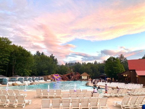 A scenic pool area with lounge chairs, a pirate ship structure, and a colorful sky at sunset surrounded by trees.