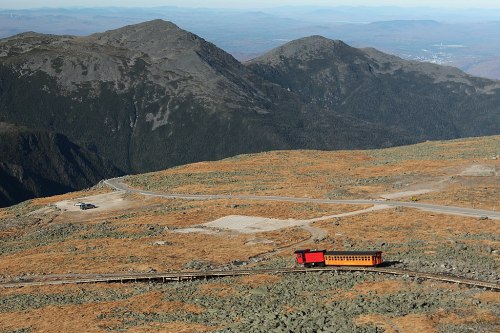 A red train travels along a winding track through a mountainous landscape with rocky terrain and distant hills.