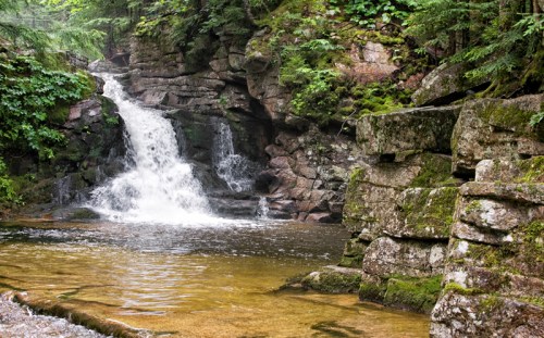 A serene waterfall cascades over rocky terrain, surrounded by lush greenery and a calm pool of water below.