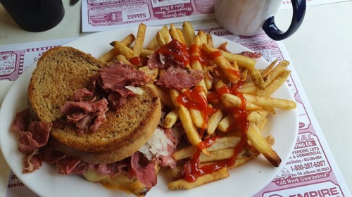 A plate featuring a sandwich with corned beef on rye bread, served with a side of fries drizzled with ketchup.