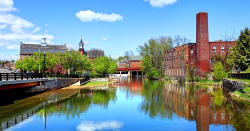 A serene river scene with green trees, blue sky, and historic buildings reflecting in the water.