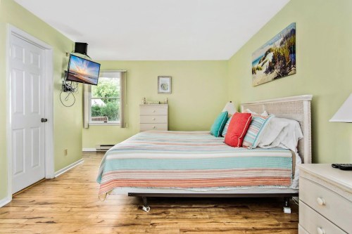 Bright bedroom with two beds, colorful pillows, a TV on the wall, and light green walls. Natural light from a window.