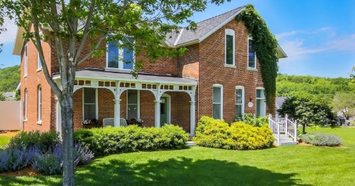 A charming brick house with a porch, surrounded by lush greenery and colorful flowers under a clear blue sky.