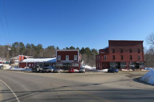 A small town scene featuring red buildings, parked cars, and a clear blue sky, with snow on the ground.