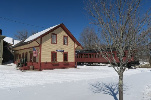 A historic train station with a red train car beside it, surrounded by snow and bare trees under a clear blue sky.