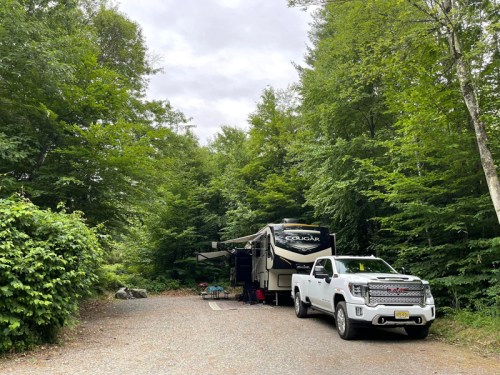 A white truck parked next to a large RV in a wooded campsite, surrounded by lush green trees and a gravel path.