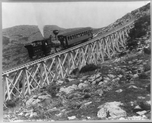 A vintage steam train travels along a steep, winding trestle track in a mountainous landscape.