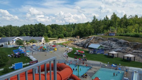 Aerial view of a water park with slides, pools, and people enjoying the outdoors surrounded by trees and blue skies.