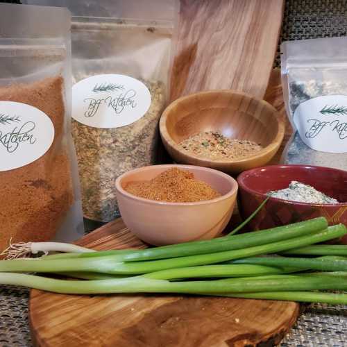 A variety of spices in labeled bags, a bowl of seasoning, and fresh green onions on a wooden surface.