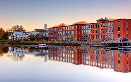 A serene waterfront scene featuring brick buildings reflecting in calm water at sunset.
