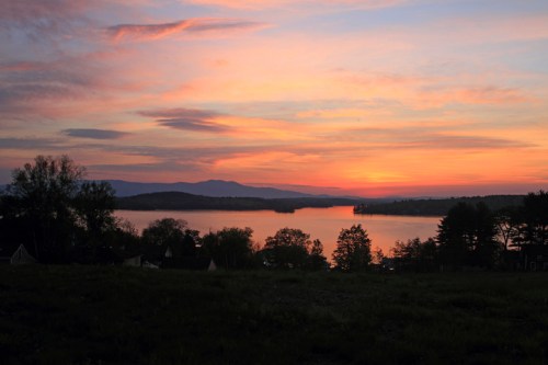 A serene sunset over a lake, with vibrant orange and pink hues reflecting on the water and silhouetted trees in the foreground.