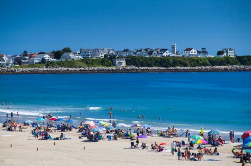 A sunny beach scene with people relaxing under umbrellas, swimming, and a row of houses in the background.