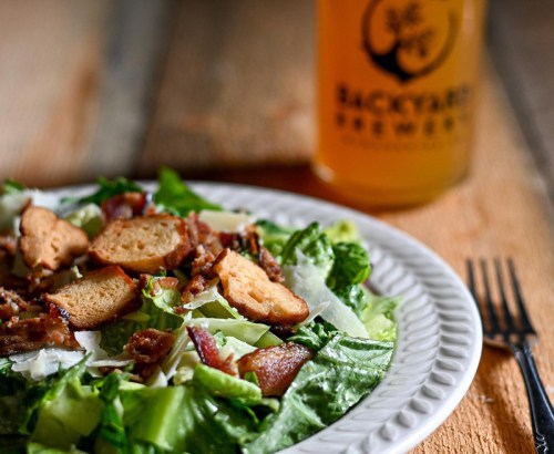 A fresh salad with bacon, croutons, and Parmesan cheese, alongside a glass of beer on a rustic wooden table.