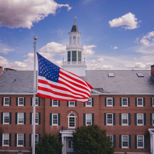 An American flag waves in front of a brick building with a clock tower under a blue sky with clouds.
