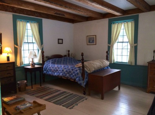Cozy bedroom with a blue quilted bed, wooden furniture, and two windows with yellow curtains.