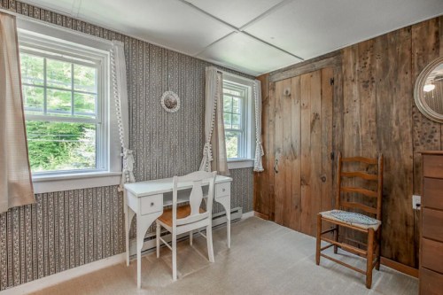 Cozy room with a white desk, wooden chair, and rustic wood paneling, featuring two windows and patterned wallpaper.