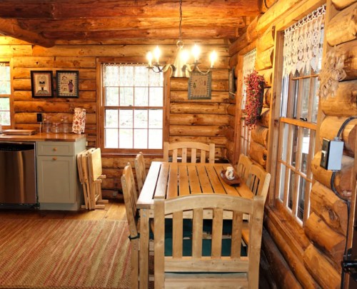 Cozy log cabin kitchen with wooden dining table, chairs, and large windows letting in natural light.