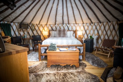Cozy yurt interior featuring a bed, wooden furniture, and warm lighting, with rugs and a rustic decor.