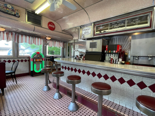 A retro diner interior featuring a counter with stools, a jukebox, and checkered flooring. Bright and inviting atmosphere.