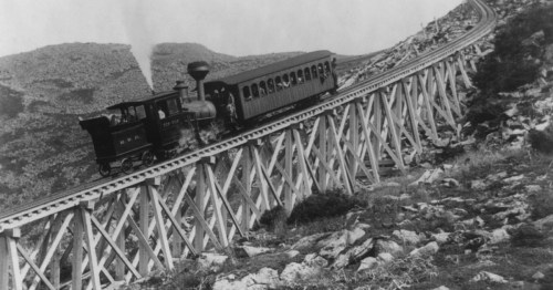 A vintage steam train traveling on a steep, wooden trestle bridge in a mountainous landscape.