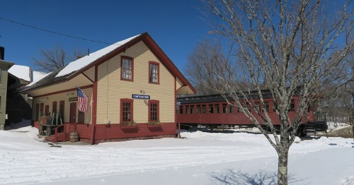 A historic train station with an American flag, surrounded by snow and a vintage train car nearby. Clear blue sky.