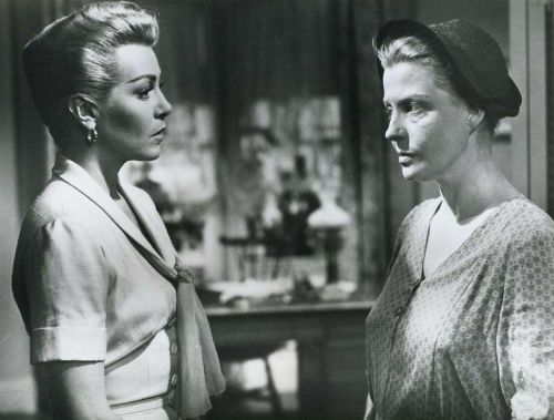 Two women in vintage clothing face each other, conveying tension, in a black-and-white setting.
