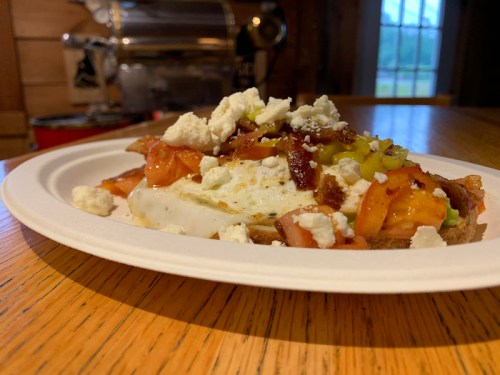 A plate of toast topped with eggs, bacon, tomatoes, pickles, and crumbled cheese, set on a wooden table.