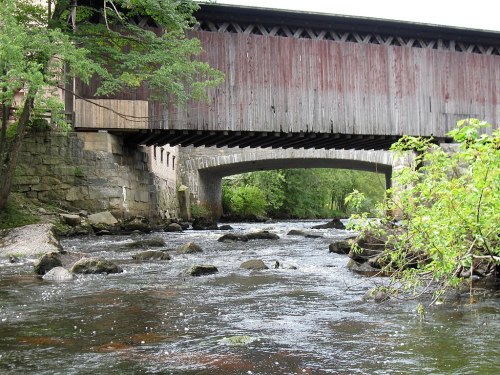A scenic view of a covered wooden bridge over a river, surrounded by greenery and rocky riverbanks.