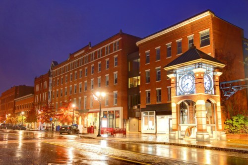 A quiet street at night featuring brick buildings and a clock tower, illuminated by streetlights after rain.