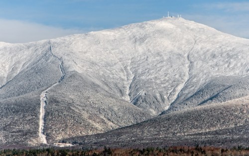 Snow-covered mountain peak with a clear sky, showcasing a mix of rocky terrain and forested slopes below.
