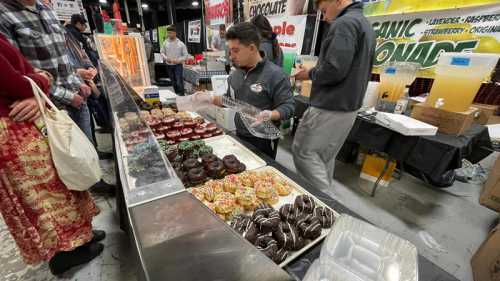 A vendor serves colorful donuts at a market, with customers browsing nearby and various food stalls in the background.