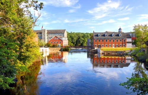 A serene river scene with historic buildings reflecting in the water, surrounded by lush greenery and a clear blue sky.