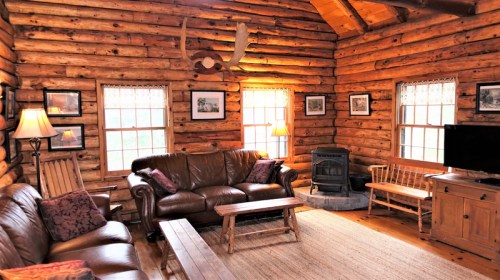 Cozy log cabin interior with leather sofas, a wood stove, and rustic decor, featuring large windows and warm lighting.