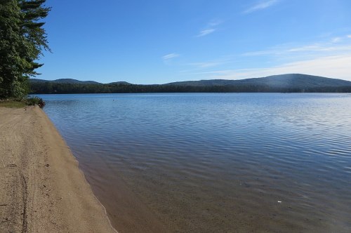 A serene lake surrounded by mountains, with a sandy shore and clear blue sky reflecting on the water's surface.