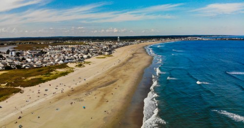 Aerial view of a sandy beach with gentle waves, bordered by a coastal town under a clear blue sky.