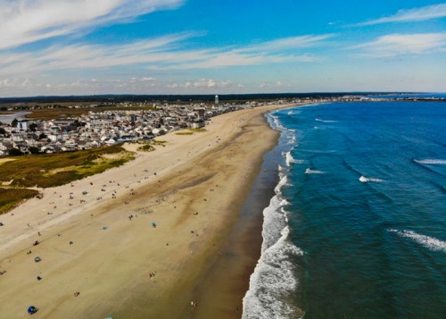 Aerial view of a sandy beach with gentle waves, coastal town in the background, and clear blue skies.